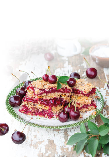 Im Mittelpunkt des Bildes steht ein Teller mit mehreren bereinander gestapelten Kirsch-Hafer-Riegeln. Die Riegel bestehen aus zwei Schichten knuspriger Hafer-Mandel-Masse, zwischen denen eine dicke, leuchtend rote Kirschf llung sichtbar ist. Die F llung wirkt saftig und leicht gl nzend, was auf frische, gut gegarte Kirschen hindeutet. Einige Kirschen, teilweise noch mit Stiel und Blatt, sind zur Dekoration auf den Riegeln sowie rund um den Teller arrangiert und unterstreichen die Frische des Geb cks.  Der Teller steht auf einem rustikalen Holztisch mit abgebl tterter, wei er Farbe, was dem Bild eine l ndlich-gem tliche, leicht nostalgische Atmosph re verleiht. Links im Hintergrund befindet sich ein Drahtkorb, prall gef llt mit frischen, dunklen Kirschen, aus dem einige Fr chte locker herausgefallen sind. Daneben steht ein kleiner wei er Emaille-Becher mit schwarzem Rand, der vermutlich Kaffee oder Espresso enth lt, sowie eine braune Holzschale mit Puderzucker im Hintergrund.  Die Szene ist hell ausgeleuchtet, mit weichem Tageslicht, das die satte rote Farbe der Kirschen und die goldbraune Kruste der Riegel besonders zur Geltung bringt. Einige gr ne Bl tter – vermutlich Kirschbl tter – erg nzen das Arrangement harmonisch.  