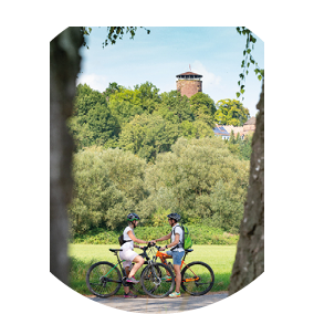 Auf dem Bild sieht man zwei Radfahrer, eine Frau und einen Mann, die sich in einer gr nen, l ndlichen Umgebung gegen berstehen. Beide tragen Helme und sportliche Kleidung, die Frau in Wei und der Mann in Grau mit kurzer Hose. Sie scheinen sich eine Pause zu g nnen und unterhalten sich, w hrend sie ihre Fahrr der nebeneinander abgestellt haben. Im Hintergrund erhebt sich ein bewaldeter H gel, auf dessen Spitze ein historischer, runder Burgturm mit einem spitzen Dach zu sehen ist. Die Szene wird von zwei Baumst mmen im Vordergrund eingerahmt, wodurch ein nat rlicher Rahmen entsteht. Die Stimmung wirkt ruhig und entspannt, ein Moment der Rast inmitten einer sommerlichen Radtour.