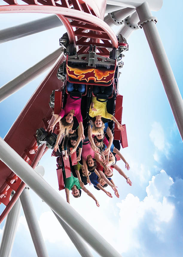 Das Bild zeigt eine Gruppe von Menschen, die auf einer roten Achterbahn sitzen, w hrend die Bahn eine enge Kurve durchf hrt und sie kopf ber h ngt. Die Personen sind sichtbar begeistert und haben die Arme nach oben gestreckt. Die Achterbahn hat mehrere Sitzreihen, die mit Sicherheitsb geln ausgestattet sind. Im Hintergrund sieht man einen strahlend blauen Himmel mit einigen wei en Wolken und die st hlernen Tr ger der Achterbahn. Die Menschen tragen bunte Kleidung, unter anderem gr n, gelb, pink und blau. Die Stimmung wirkt fr hlich und aufregend.   M chtest du die Beschreibung noch ausf hr