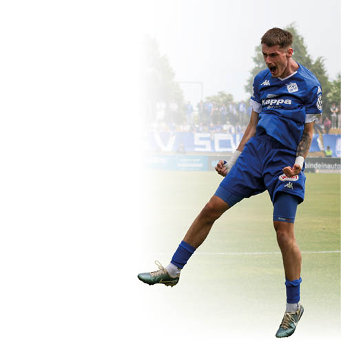 Auf dem Bild ist ein junger m nnlicher Fu ballspieler in einem blauen Trikot zu sehen. Er befindet sich auf einem Fu ballfeld und scheint gerade einen Torerfolg gefeiert zu haben. Der Spieler springt leicht in die Luft, seine F uste sind angespannt und er hat den Mund weit ge ffnet, als w rde er jubeln oder laut schreien. Er tr gt kurze dunkelblonde Haare und an beiden H nden sind seine Handgelenke bandagiert. Im Hintergrund ist ein weiterer Spieler in hnlicher Kleidung zu sehen, der l chelnd in Richtung des jubelnden Spielers l uft. Im Hintergrund befinden sich au erdem einige Zuschauer und B ume, sowie Werbebanden am Spielfeldrand. Das Wetter wirkt bew lkt, die Atmosph re ist lebendig und sportlich.