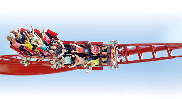 Das Bild zeigt eine Achterbahn in voller Fahrt. Es handelt sich um einen roten Zug mit zwei Reihen an Sitzen, der sich gerade in einer berkopf-Schleife (Looping) befindet. Der Zug ist kopf ber – die Passagiere h ngen also mit den K pfen nach unten in der Luft.    Die Mitfahrenden,  berwiegend Jugendliche, tragen bunte Sommerkleidung in Farben wie Gelb, Rosa, T rkis und Schwarz. Viele haben die Arme in die Luft gestreckt und ihre Haare wehen durch die starke Bewegung nach unten. Ihre Gesichtsausdr cke zeigen Freude, Aufregung oder auch Schrecken – typisch f r eine rasante Achterbahnfahrt.    Die Schienen der Achterbahn sind knallrot, ebenso wie die Sicherheitsb gel und Sitzr cklehnen. Der Hintergrund ist ein wolkenloser Himmel in zartem Blau mit einem leichten Verlauf nach Wei  oben – was die Geschwindigkeit und H he der Szene betont.    Das Bild vermittelt ein Gef hl von Adrenalin, Nervenkitzel und Freizeitspa .
