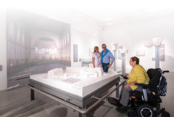 In einem hellen Museumsraum steht ein gro er rechteckiger Tisch mit einem wei en Architekturmodell einer historischen Geb udeanlage. Links an der Wand h ngt ein gro formatiges Bild, das den Innenraum einer Halle mit hohen Fenstern und einer gew lbten Decke zeigt. Rechts im Bild sitzt eine Frau im elektrischen Rollstuhl, sie tr gt eine gelbe Strickjacke und graue Hose. Sie schaut interessiert auf das Modell. Auf der anderen Seite des Tisches stehen ein Mann und eine Frau nebeneinander, beide l cheln und blicken ebenfalls auf das Modell. Im Hintergrund sind auf Podesten mehrere wei e steinerne S ulenkapitelle und andere historische Bruchst cke ausgestellt. Die Szene vermittelt den Eindruck eines inklusiven und zug nglichen Museumsbesuchs.