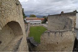 Die Aufnahme zeigt einen Innenhof, umgeben von hohen, dicken Mauern aus grob behauenen, hellen Steinen. Links im Bild ist ein gro er gemauerter Bogen in der Mauer zu sehen. Der Boden des Hofs ist mit gepflegtem gr nen Rasen bedeckt. Im Hintergrund stehen moderne Geb ude mit roten und grauen D chern, dahinter sind B ume und eine leicht h gelige Landschaft zu erkennen. Der Himmel ist mit grauen Wolken bedeckt, was der Szene ein eher k hles und ged mpftes Licht verleiht. Die Mauern wirken wie berreste einer historischen Befestigungsanlage oder eines alten Bauwerks.