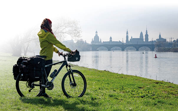 Eine Person steht mit einem vollbepackten Reiserad auf einer gr nen Wiese am Ufer eines breiten Flusses. Sie tr gt eine leuchtend gelbe Regenjacke und ein rotes Tuch ber den Haaren. Am Fahrrad sind gro e schwarze Packtaschen befestigt, sowohl hinten als auch vorne. Die Person schaut nach rechts  ber den Fluss, wo eine steinerne Br cke mit mehreren B gen zu sehen ist. Dahinter erhebt sich die Silhouette einer Stadt mit vielen T rmen und Kuppeln, teilweise im Dunst verschwommen. Der Himmel ist hell und leicht neblig, was der Szene eine ruhige, fast vertr umte Stimmung gibt.