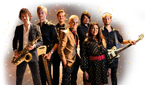 Das Bild zeigt sieben Menschen, die vor einem dunklen, mit goldenen Lichtpunkten durchzogenen Hintergrund posieren. Sie sind alle elegant gekleidet und wirken wie eine Musikgruppe. Ganz links steht ein Mann mit lockigem braunem Haar, der einen schwarzen Anzug mit wei em Hemd tr gt und ein Saxophon h lt. Neben ihm steht ein Mann mit kurzen blonden Haaren, schwarzem Hemd und einer Bassgitarre. Dritter von links ist ein Mann mit kurzem, hellblondem Haar, der einen goldfarbenen Blazer, ein rotes Hemd und eine gestreifte Hose tr gt. Hinter ihm, leicht verdeckt, steht ein Mann mit mittellangem dunklem Haar und schwarzem Hemd. Rechts vorne steht eine Frau mit langen, dunklen Haaren, die ein schwarzes Kleid mit goldfarbenen Mustern und einen auff lligen roten G rtel tr gt. Ganz rechts h lt ein Mann mit kurzem blonden Haar und schwarzem Hemd eine E-Gitarre. Alle Personen schauen direkt in die Kamera und wirken freundlich und selbstbewusst.