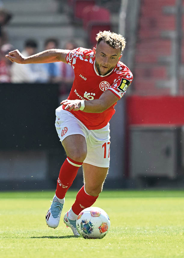 Auf dem Bild ist ein Fu ballspieler in Aktion zu sehen. Er tr gt ein rotes Trikot mit wei en Mustern auf den Schultern und wei e Hosen. Auf dem Trikot ist das Logo vom Verein Mainz 05. Der Spieler l uft schnell und schaut konzentriert auf den Ball, den er gerade mit dem Fu f hrt. Im Hintergrund sieht man unscharf Zuschauer auf der Trib ne. Die Sonne scheint – es ist ein Spiel bei Tageslicht.