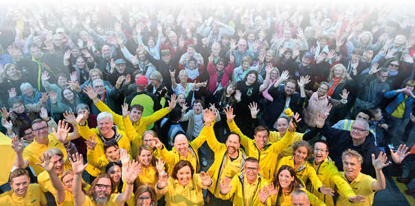 Das Bild zeigt eine gro e fr hliche Menschenmenge, die in die Kamera winkt. Vorne stehen viele Personen in gelben Jacken oder Shirts – ein Team vom SWR. Dahinter sieht man viele weitere Menschen, dicht gedr ngt, bunt gekleidet. Alle wirken gut gelaunt. Die Stimmung ist lebendig und freundlich – wie bei einem Fest oder einer ffentlichen Veranstaltung.