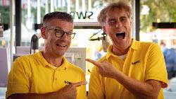 Auf dem Bild sind zwei M nner zu sehen, die fr hlich lachen. Beide tragen gelbe Poloshirts mit dem Logo vom SWR (S dwestrundfunk). Sie stehen nebeneinander in einem hellen Raum mit viel Tageslicht. Beide zeigen mit dem Finger auf den jeweils anderen und wirken gut gelaunt. Im Hintergrund sieht man unscharf Menschen und etwas Technik. Die Stimmung ist locker und freundlich.