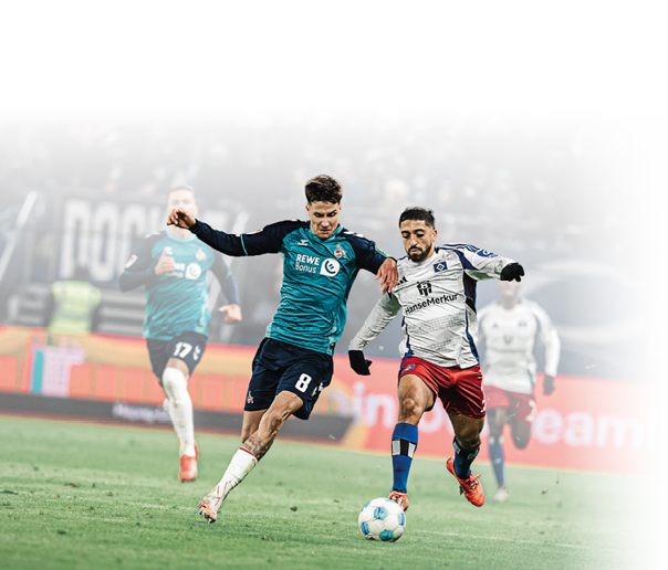 Auf dem Bild sind zwei Fu ballspieler im Zweikampf um den Ball zu sehen. Der Spieler links tr gt ein blau-t rkisfarbenes Trikot mit dem Logo des 1. FC K ln und der Aufschrift „REWE Bonus“. Der Spieler rechts tr gt ein wei es Trikot mit roten Hosen – das ist der Hamburger SV. Beide Spieler sind in Bewegung und k mpfen energisch um den Ball, der zwischen ihnen liegt. Im Hintergrund sieht man weitere Spieler im Stadion. Die Szene ist dynamisch und zeigt den Einsatz beider Spieler im Spiel.