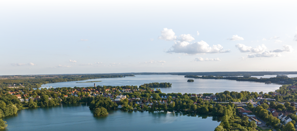 Das Bild zeigt eine wundersch ne Luftaufnahme einer Stadt an mehreren Seen. Die Landschaft ist gr n und friedlich, mit vielen B umen und kleinen H usern. In der Mitte liegt eine Halbinsel mit dicht bebautem Ortskern. Mehrere Seen erstrecken sich in alle Richtungen – das Wasser ist ruhig und blau. Am Horizont sieht man W lder und weite Landschaften. Der Himmel ist klar mit ein paar wei en Wolken. Die Szenerie wirkt sommerlich und idyllisch – ideal zum Entspannen, Spazieren oder Bootfahren.