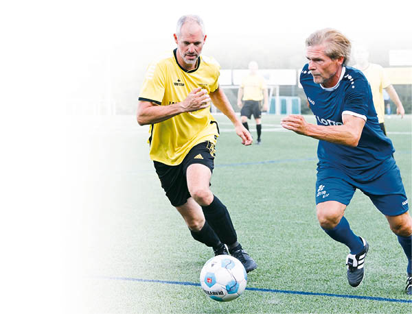 Das Bild zeigt eine spannende Fu ballszene auf einem Kunstrasenplatz. Zwei Spieler liefern sich ein intensives Laufduell um den Ball. Der Spieler links tr gt ein gelb-schwarzes Trikot. Der Spieler rechts tr gt ein blaues Trikot mit der Aufschrift „Lotto“. Beide schauen konzentriert auf den Ball und sind in vollem Sprint.