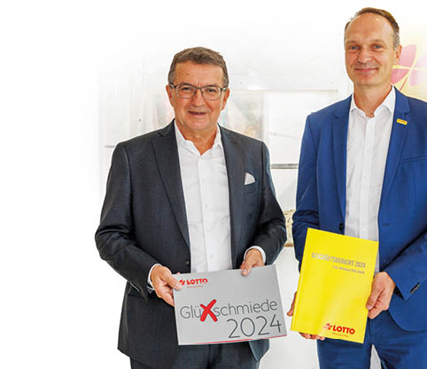 Auf dem Bild sind zwei M nner in Anz gen zu sehen, die nebeneinanderstehen und freundlich in die Kamera blicken. Der Mann links h lt eine Mappe mit der Aufschrift „Gl cksschmiede 2024“ und dem LOTTO Rheinland-Pfalz-Logo. Der Mann rechts h lt den Gesch ftsbericht 2024 von LOTTO Rheinland-Pfalz in der Hand. Im Hintergrund befindet sich ein gelber Aufsteller mit dem bekannten roten Lotto-Kleeblatt-Logo. Das Bild vermittelt einen offiziellen, professionellen Anlass – es handelt sich um die Pressekonferenz zur Jahresbilanz von Lotto Rheinland-Pfalz
