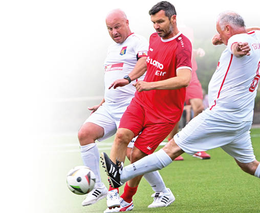 as Bild zeigt eine intensive Spielsituation auf dem Fu ballfeld:   Drei  ltere Spieler k mpfen energisch um den Ball. Der Spieler in der Mitte tr gt das rote Trikot der LOTTO Elf.    Die beiden Spieler in Wei  – mit dem Wappen des Gegners auf der Brust – setzen zur Gr tsche oder zum Block an.    Alle drei sind hochkonzentriert und k rperlich engagiert im Zweikampf.    Die Szene symbolisiert fairen, ambitionierten Altherren-Fu ball mit sportlichem Ehrgeiz. Das Bild ist dynamisch, sportlich und zeigt, dass Fu ballbegeisterung keine Altersgrenze kennt.