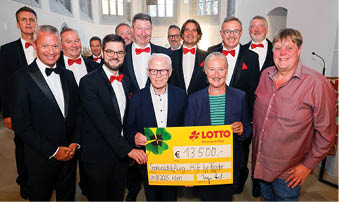 Auf dem Foto sind die Mainzer Hofs nger in der evangelischen Kirche in Kirn zu sehen. Die M nner des Chores tragen schwarze Anz ge mit wei en Hemden und roten Fliegen. In ihrer Mitte stehen drei Personen mit einem gro en gelben Spendenscheck von Lotto Rheinland-Pfalz ber 13.500 Euro. Von links nach rechts: ein Chorvertreter, Stiftungs-Vorsitzender Herbert Wirzius mit wei em Haar und Brille, Lotto-Gesellschaftervertreterin Monika Sauer im dunkelblauen Blazer und daneben Stadtb rgermeister Frank Ensminger im roten Hemd. Alle l cheln in die Kamera, im Hintergrund ist der helle Kirchenraum mit hohen Fenstern zu erkennen.