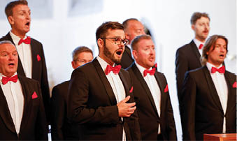 Auf dem Foto sind die Mainzer Hofs nger in der evangelischen Kirche in Kirn zu sehen. Die M nner des Chores tragen schwarze Anz ge mit wei en Hemden und roten Fliegen. In ihrer Mitte stehen drei Personen mit einem gro en gelben Spendenscheck von Lotto Rheinland-Pfalz ber 13.500 Euro. Von links nach rechts: ein Chorvertreter, Stiftungs-Vorsitzender Herbert Wirzius mit wei em Haar und Brille, Lotto-Gesellschaftervertreterin Monika Sauer im dunkelblauen Blazer und daneben Stadtb rgermeister Frank Ensminger im roten Hemd. Alle l cheln in die Kamera, im Hintergrund ist der helle Kirchenraum mit hohen Fenstern zu erkennen.