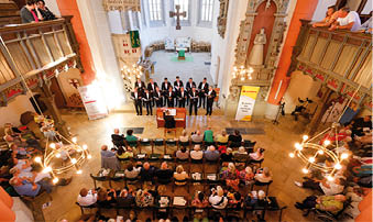 Auf dem Foto sind die Mainzer Hofs nger in der evangelischen Kirche in Kirn zu sehen. Die M nner des Chores tragen schwarze Anz ge mit wei en Hemden und roten Fliegen. In ihrer Mitte stehen drei Personen mit einem gro en gelben Spendenscheck von Lotto Rheinland-Pfalz ber 13.500 Euro. Von links nach rechts: ein Chorvertreter, Stiftungs-Vorsitzender Herbert Wirzius mit wei em Haar und Brille, Lotto-Gesellschaftervertreterin Monika Sauer im dunkelblauen Blazer und daneben Stadtb rgermeister Frank Ensminger im roten Hemd. Alle l cheln in die Kamera, im Hintergrund ist der helle Kirchenraum mit hohen Fenstern zu erkennen.