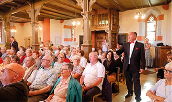 Auf dem Foto sind die Mainzer Hofs nger in der evangelischen Kirche in Kirn zu sehen. Die M nner des Chores tragen schwarze Anz ge mit wei en Hemden und roten Fliegen. In ihrer Mitte stehen drei Personen mit einem gro en gelben Spendenscheck von Lotto Rheinland-Pfalz ber 13.500 Euro. Von links nach rechts: ein Chorvertreter, Stiftungs-Vorsitzender Herbert Wirzius mit wei em Haar und Brille, Lotto-Gesellschaftervertreterin Monika Sauer im dunkelblauen Blazer und daneben Stadtb rgermeister Frank Ensminger im roten Hemd. Alle l cheln in die Kamera, im Hintergrund ist der helle Kirchenraum mit hohen Fenstern zu erkennen.