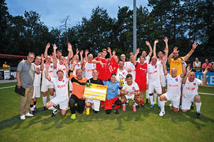 Das Bild zeigt eine gro e Gruppe fr hlicher Menschen auf einem Sportplatz. Sie haben sich zur bergabe eines symbolischen Spendenschecks von 10.000 € versammelt, gestiftet von LOTTO Rheinland-Pfalz.    Im Vordergrund steht ein gelbes „LOTTO Elf“-Schild. In der Mitte wird der Scheck hochgehalten – die Spende geht an ein Projekt zur Unterst tzung von Waisenkindern in Ruanda, wie aus der Aufschrift hervorgeht.    Die Gruppe besteht aus Sportlern (erkennbar an den roten „LOTTO Elf“-Trikots), Organisatoren, Unterst tzern und G sten – teils in Alltagskleidung, teils in thematisch passenden Shirts (z. B. „Rwanda“). Die Stimmung ist herzlich, engagiert und gemeinschaftlich – ein starkes Zeichen f r soziales Engagement durch Sport.