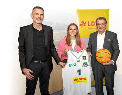 Drei Personen stehen nebeneinander in einem Raum mit Holzw nden und einem gro en Foto der Festung Ehrenbreitstein im Hintergrund. Links steht Thomas Klein im schwarzen Anzug, in der Mitte Emilia Keller im pinkfarbenen Blazer, die ein wei es Basketballtrikot der EPG Baskets mit Lotto-Logo h lt. Rechts steht J rgen H fner im grauen Anzug und h lt einen Basketball in den H nden. Neben ihm ist ein gelbes Roll-up mit dem Lotto-Logo zu sehen.