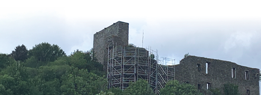 Die Fotografie zeigt die Ruine der Virneburg in der Eifel. Sie liegt auf einem bewaldeten H gel. Links im Bild erhebt sich ein quadratischer Burgturm, an dessen Seite ein gro es Bauger st angebracht ist. Dieses mehrst ckige Ger st verdeckt teilweise die Mauer und deutet auf Sanierungs- oder Sicherungsarbeiten hin. Rechts daneben ist ein l nglicher Geb udeteil zu sehen, dessen graue Steinmauern noch stehen. Fenster- und T r ffnungen sind gut erkennbar, ein Dach fehlt. Dichtes, gr nes Laub umgibt die Ruine. Am Himmel h ngen graue Wolken, es wirkt leicht tr b und regnerisch.