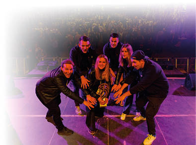 Sechs Mitglieder der Band HOTSPOT posieren auf einer B hne. Sie stehen im Halbkreis um die Troph e, die eine Frau in der Mitte h lt. Alle strecken die H nde jubelnd in Richtung Pokal. Im Hintergrund ist das Publikum vor der B hne zu sehen, die Szene ist in violettes B hnenlicht getaucht.