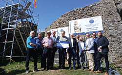 Vor einer Burgruine mit Bauger st und Banner der Deutschen Stiftung Denkmalschutz steht eine Gruppe von zw lf Personen. Zwei M nner in der Mitte halten symbolisch einen F rdervertrag. Hinter ihnen ist die alte Bruchsteinmauer der Burg zu sehen, dar ber strahlt blauer Himmel.