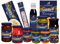 Das Bild zeigt eine Auswahl italienischer Lebensmittel der Marke „Leverno“. Die Produkte sind in Gl sern, Flaschen, Dosen und Kartons verpackt. Sichtbar sind unter anderem Grissini in einer blauen Schachtel, Torrone (eine wei e Nougatspezialit t mit Mandeln) in einer langen Verpackung, eine Flasche Balsamico-Creme, Lasagnebl tter in einer blauen Schachtel sowie verschiedene eingelegte und verarbeitete Produkte im Glas. Dazu geh ren getrocknete Tomaten, gegrillte Paprika, Pesto in unterschiedlichen Sorten, eine Gem secreme, eine Tomatenso e und ein Glas mit eingelegten gr nen Bohnen. Alle Produkte tragen ein dunkelblaues Etikett mit goldfarbenem Schriftzug und L wen-Logo der Marke „Leverno“.