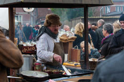 Das Foto zeigt einen Mann an einem Marktstand, der Speisen in einer gro en Pfanne zubereitet. Er tr gt eine braune Fellm tze mit Ohrenklappen, einen dicken Schal, ein gestreiftes Hemd und eine braune Ledersch rze. Neben ihm liegen verschiedene Lebensmittel, darunter Kartoffeln, Brotlaibe in einem Korb und Konservendosen. Im Hintergrund stehen viele Menschen, die den Markt besuchen. Vor dem Stand ist auch ein M dchen mit blondem Zopf zu sehen, das neugierig in Richtung der Pfanne schaut. Aus der Pfanne steigt Dampf auf, was eine winterlich-k hle Atmosph re vermittelt.