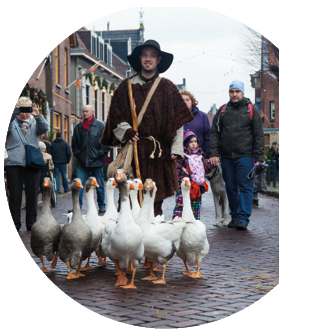Das Foto zeigt eine Stra enszene mit einem Mann, der eine Gruppe von G nsen anf hrt. Er tr gt ein braunes, grobes Gewand, einen breiten Hut und eine Umh ngetasche – er wirkt wie ein G nsehirt in historischer Kleidung. Vor ihm laufen etwa zehn G nse dicht zusammen ber das Kopfsteinpflaster. Im Hintergrund stehen Menschen am Stra enrand, darunter Erwachsene und Kinder, die die Szene beobachten. Einige tragen Winterjacken, es scheint k hl zu sein. Die Stra e ist von H usern mit weihnachtlicher Dekoration ges umt.