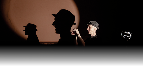 Das Foto zeigt einen Mann mit grauem Hut vor einer Wand, auf die ein starkes Licht f llt. Durch das Licht wird ein gro er Schatten seines Kopfes und Oberk rpers auf die Wand geworfen. Der Schatten wirkt bergro  und deutlich erkennbar, w hrend der Mann selbst rechts im Bild steht und l chelt. Er h lt die Hand erhoben, als w rde er mit seinem Schatten spielen oder eine Geste machen. Links auf der Wand ist zus tzlich ein kleinerer Schattensilhouettenkopf mit Hut zu sehen, sodass es aussieht, als w rden die Schattenfiguren miteinander interagieren.