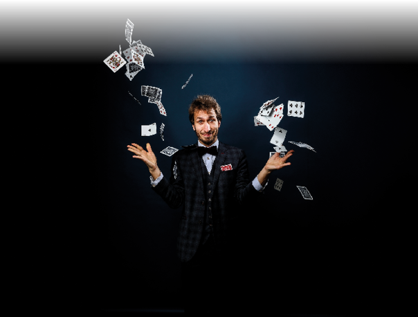 Das Foto zeigt einen Mann in einem dunklen, karierten Anzug mit Fliege, der vor einem dunklen Hintergrund steht. Er l chelt verschmitzt und h lt beide Arme seitlich nach au en. Um ihn herum fliegen zahlreiche Spielkarten durch die Luft, als h tte er sie gerade hochgeworfen. In seiner Brusttasche steckt ebenfalls eine Spielkarte. Die Szene wirkt wie ein magischer Moment, passend zu einem Zaubertrick oder einer Jonglage mit Karten.