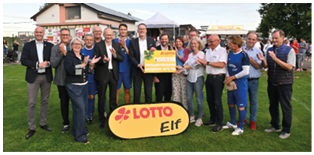 Das Foto zeigt eine Gruppe von etwa 15 Personen, die gemeinsam auf einem Fu ballfeld stehen. In der Mitte wird ein gro er symbolischer Scheck in H he von 25.000 Euro pr sentiert, der von „LOTTO Rheinland-Pfalz“ stammt. Mehrere Personen klatschen und l cheln, die Stimmung wirkt feierlich. Einige der Anwesenden tragen blaue Fu balltrikots mit der Aufschrift „LOTTO Elf“, andere sind in Alltagskleidung oder Anz gen gekleidet. Vor der Gruppe steht ein gelbes Schild mit dem roten LOTTO-Logo und der Aufschrift „Elf“. Im Hintergrund sind ein Vereinsheim, Zuschauer sowie weitere Menschen zu sehen. 