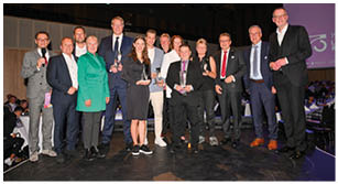 Das Foto zeigt eine Gruppe von rund 14 Personen auf einer B hne bei einer Feier zum 75. Jubil um des Landessportbundes Rheinland-Pfalz. Links steht ein Mann im grauen Anzug mit Mikrofon, der offenbar moderiert. Mehrere Personen halten Glastroph en in den H nden – es handelt sich um eine Preisverleihung. Die Anwesenden tragen unterschiedliche Kleidung, von eleganten Anz gen und Kleidern bis zu sportlich-legeren Outfits. Im Hintergrund sind festlich gedeckte Tische, G ste und eine gro e Leinwand mit dem Jubil umslogo „75 Jahre LSB Rheinland-Pfalz“ zu sehen. Die Stimmung wirkt feierlich 