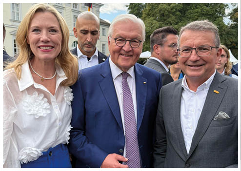 Eine Frau und zwei M nner l cheln in die Kamera. Im Hintergrund sind ein Schloss sowie mehrere Personen zu erkennen.