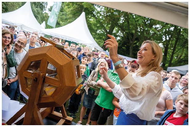 Eine elegant gekleidete Frau steht auf einer B hne an einer Lostrommel. Vor der B hne schauen viele Menschen gespannt der Verlosung zu. Im Hintergrund sind Pavillonzelte zu erkennen. 