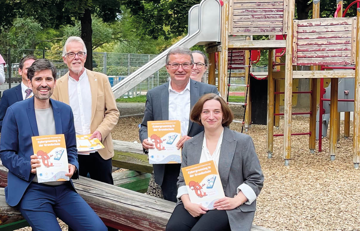 Das Foto zeigt eine Gruppe von Kindern und Erwachsenen auf einem Spielplatz. Die Kinder sitzen und stehen auf einer gro en, holzgeschnitzten Wippe in Form eines Bleistifts. Sie lachen, winken oder halten bunte Geschenke in den H nden. Die Erwachsenen – M nner im Anzug und eine Frau im Blazer – sitzen ebenfalls auf der Wippe und halten orangefarbene Brosch ren mit der Aufschrift „Fit f rs Handy“ in die Kamera. Im Hintergrund sind B ume sowie ein Kletterger st mit Rutsche zu sehen.