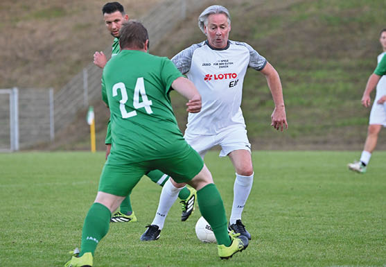 Auf einem Fu ballfeld spielt ein Mann in wei em Trikot der „LOTTO Elf“ mit dem Ball. Er l uft auf einen Gegenspieler in gr nem Trikot mit der R ckennummer 24 zu, der ihn zu stoppen versucht. Im Hintergrund sind weitere Spieler zu sehen, teils in gr ner, teils in wei er Spielkleidung. Die Szene wirkt dynamisch und zeigt einen spannenden Moment w hrend des Spiels.