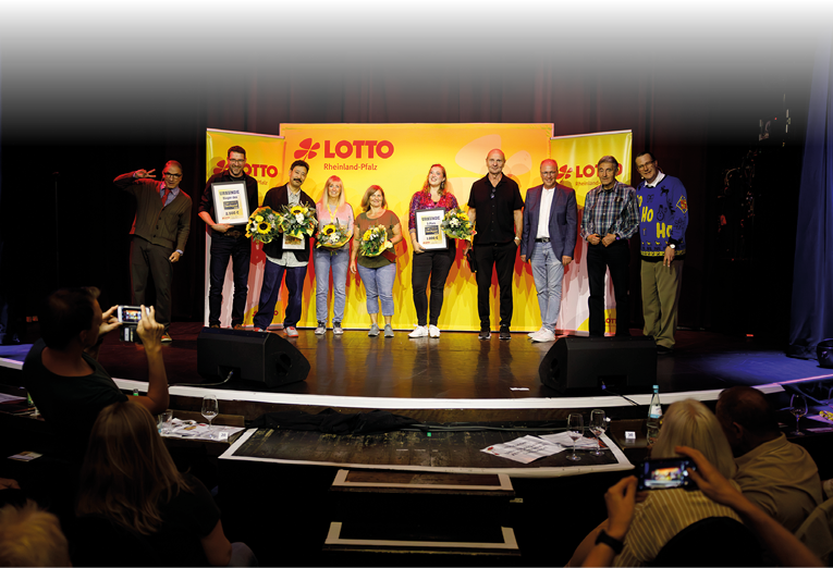 Auf dem Bild ist eine Gruppe von zehn Personen auf einer B hne zu sehen. Sie stehen vor einem gelben Hintergrund mit dem roten Logo von „LOTTO Rheinland-Pfalz“. Die Stimmung wirkt feierlich und fr hlich, einige Personen halten Urkunden und Blumenstr u e in den H nden.