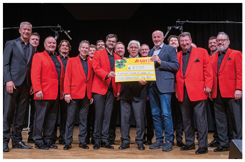 Eine gr ere Gruppe von M nnern steht in zwei Reihen auf einer B hne. Die meisten tragen schwarze Hosen, schwarze Hemden und rote Sakkos, einige wenige tragen dunkle Anz ge. In der Mitte halten zwei M nner einen gro en gelben symbolischen Scheck, auf dem eine Spendensumme von 8.500 Euro zu sehen ist. Im Hintergrund h ngt ein schwarzer Vorhang, dar ber sind Mikrofone an St ndern zu erkennen. Der Boden ist aus Holz.