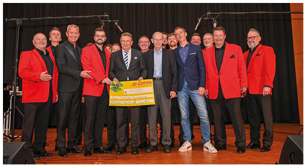 Eine Gruppe von M nnern steht auf einer B hne vor einem schwarzen Vorhang. Einige tragen rote Sakkos mit schwarzer Kleidung, andere dunkle Anz ge oder ein Sakko mit Jeans. In der Mitte halten mehrere Personen gemeinsam einen gro en gelben symbolischen Scheck. ber der Gruppe h ngen zwei Mikrofone an langen St ndern von oben herab. Der Boden der B hne ist aus Holz.