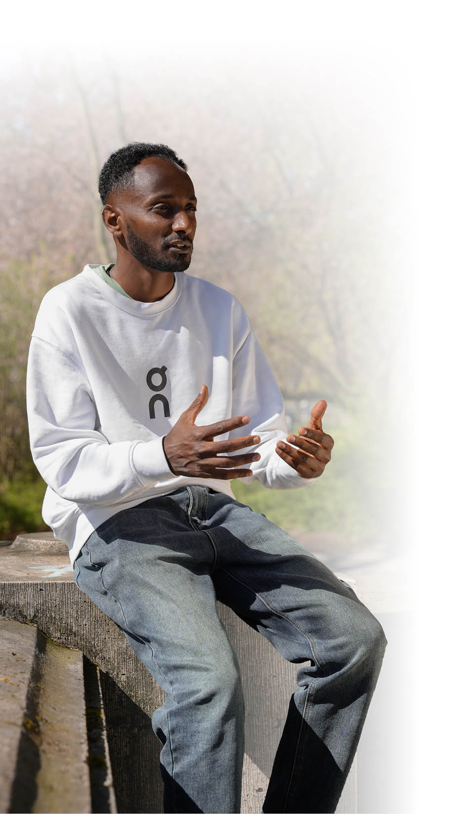 Das Bild zeigt einen jungen Mann mit kurzem, dunklem, lockigem Haar und einem kurzen, gepflegten Bart. Er sitzt auf einer niedrigen Betonmauer im Freien, vermutlich in einem Park. Der Mann tr gt ein wei es Sweatshirt mit einem stilisierten, senkrechten Schriftzug „on“ auf der Vorderseite sowie eine locker sitzende, blaue Jeans.   Er wirkt konzentriert und spricht gestikulierend mit beiden H nden, wobei seine Finger leicht gespreizt sind. Seine K rperhaltung ist aufrecht, er schaut aufmerksam in Richtung einer Person oder Kamera au erhalb des Bildausschnitts.    Im Hintergrund sind gr n-braune B ume zu sehen, die teils schon Bl tter tragen, was auf Fr hling hindeutet. Die Sonne scheint, es ist Tageslicht, und das Wetter wirkt angenehm.   