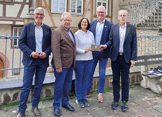 F nf Personen stehen nebeneinander im Kauber Ortskern mit Fachwerkh usern. Zwei M nner tragen dunkle Anz ge, einer davon h lt eine Plakette gemeinsam mit einer Frau im wei en Pullover. Die Gruppe l chelt in die Kamera, im Hintergrund sind die detailreichen Fachwerkfassaden und eine Steintreppe zu sehen. 