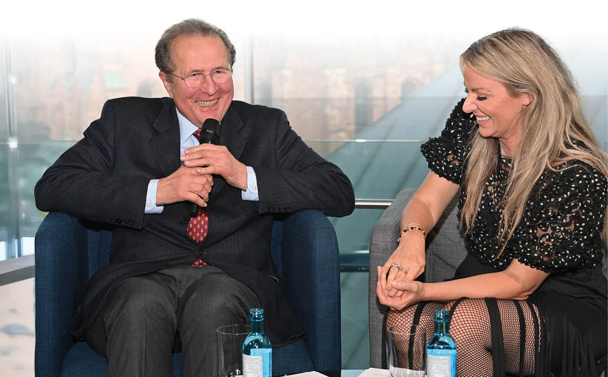 Ein lterer Herr im Anzug mit roter Krawatte sitzt l chelnd auf einem blauen Sessel und h lt ein Mikrofon in der Hand. Neben ihm sitzt eine blonde Frau im schwarzen Kleid, die ebenfalls lacht. Vor ihnen steht ein Glastisch mit Wasserflaschen und Gl sern. Im Hintergrund ist durch gro e Fenster schemenhaft die Fassade des Mainzer Doms sichtbar.