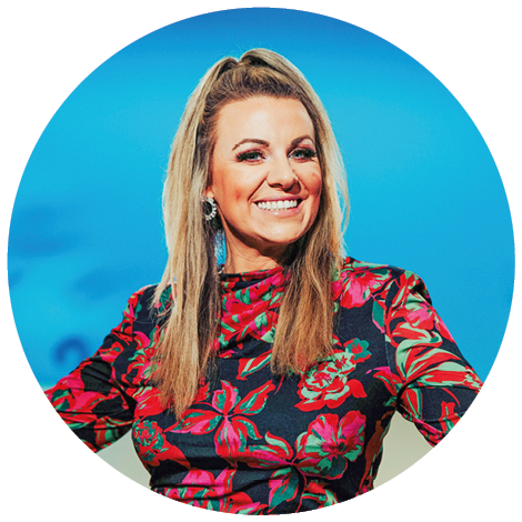 Das Bild zeigt eine l chelnde Frau vor einem blauen Hintergrund – sie tr gt ein farbenfrohes, gebl mtes Oberteil und wirkt sympathisch und pr sentationsfreudig.