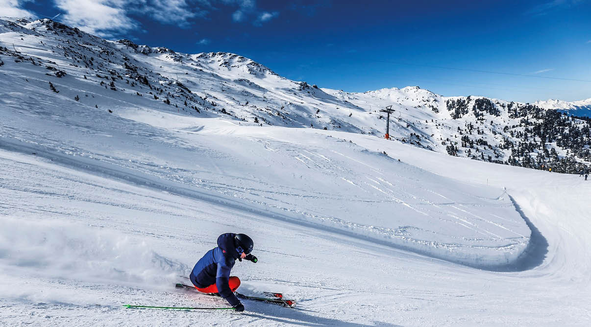 Das Bild zeigt eine Person, die auf einer schneebedeckten Piste Ski f hrt. Die Person tr gt eine blaue Jacke, rote Skihosen, schwarze Handschuhe und einen schwarzen Helm. In dynamischer Kurvenlage zieht sie eine enge Spur durch den Schnee, wobei feiner Pulverschnee zur Seite spritzt. Im Hintergrund breiten sich verschneite Berggipfel unter einem strahlend blauen Himmel aus. Die Szene vermittelt Bewegung, sportliche Energie und winterliche Frische.