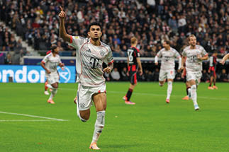 Das Bild zeigt den Fu ballspieler Luis D az vom FC Bayern M nchen, der offenbar gerade ein Tor gefeiert. Er tr gt das wei e Ausw rtstrikot mit grauen und roten Akzenten und hebt den rechten Arm zum Jubel. Im Hintergrund sind Spieler der eigenen und der gegnerischen Mannschaft – in schwarz-roten Trikots – sowie zahlreiche Zuschauer im Stadion zu sehen.