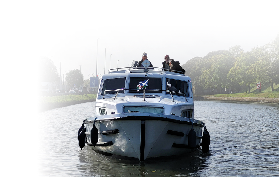 Das Bild zeigt ein Hausboot, das ruhig auf einem Kanal unterwegs ist. Drei Personen stehen auf dem Deck und genie en die Fahrt – die Sonne scheint, das Wasser ist still, und am Ufer sind B ume im sanften Morgen- oder Abendlicht zu erkennen.   Die Perspektive direkt von vorn verleiht der Aufnahme Dynamik – es wirkt fast so, als w rde das Boot direkt auf den Betrachter zufahren. Die schwarzen Fender an der Seite und die klaren Linien des Bootsrumpfes betonen den robusten, funktionalen Stil moderner Hausboote.