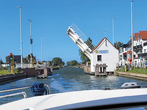 Das Bild zeigt eine idyllische Szene auf einem Kanal – offenbar aus der Perspektive eines Bootes. Im Vordergrund ist der Bug des Bootes zu sehen, w hrend sich im Hintergrund eine ge ffnete Zugbr cke ber dem Wasser erhebt. Das kleine wei e Br ckenh uschen mit dem Schild „Lutjegastbrug“ und die umliegenden H user deuten darauf hin, dass die Aufnahme in Flandern entstanden ist.    Der Himmel ist klarblau, das Licht hell – es wirkt wie ein sonniger Sommertag auf einer gem tlichen Bootstour durch die friesische Landschaft.