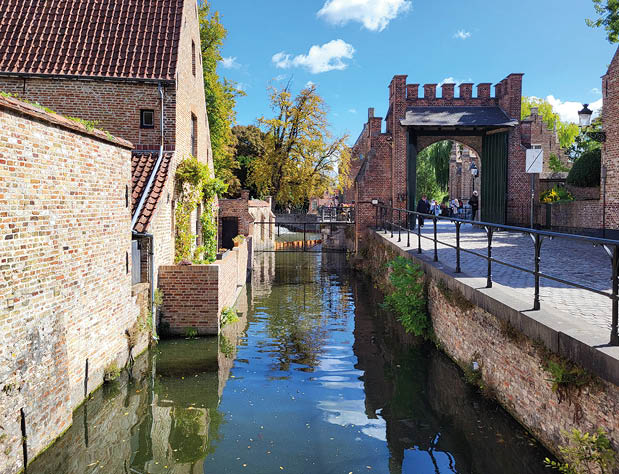 Das Bild zeigt den idyllischen Beginenhof von Br gge, eine der bekanntesten historischen St tten der belgischen Stadt. Zu sehen ist ein schmaler, von alten Backsteinmauern ges umter Kanal, der auf das markante Torhaus zuf hrt. Das Licht des Sp tsommers spiegelt sich auf dem Wasser, w hrend Spazierg nger durch das Tor in die friedliche Anlage eintreten.