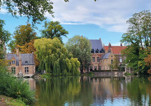 Das Foto zeigt eine idyllische, fast m rchenhafte Szene mit historischen Geb uden an einem stillen Gew sser. Im Vordergrund spiegeln sich die H user und ppigen B ume – darunter eine prachtvolle Trauerweide – im ruhigen Wasser. Die Architektur mit ihren Backsteinfassaden und spitzen Giebeln erinnert stark an fl mische Baukunst, typisch f r St dte wie Br gge in Belgien.    Das Licht und die Farben deuten auf einen milden Tag im Fr hherbst hin, mit goldenem Laub und noch sattem Gr n – eine friedliche, malerische Stimmung.